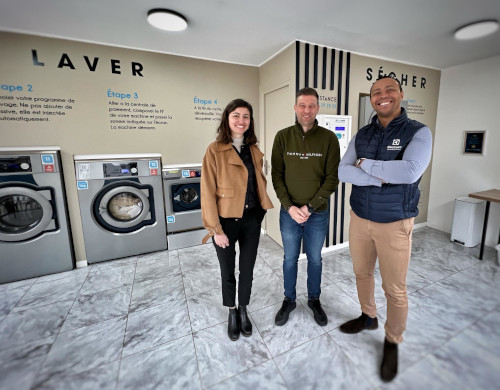 why_Laverie de Saint Omer Three people, smiling, in the center of a new laundry shop, with washing machines and a dryer.